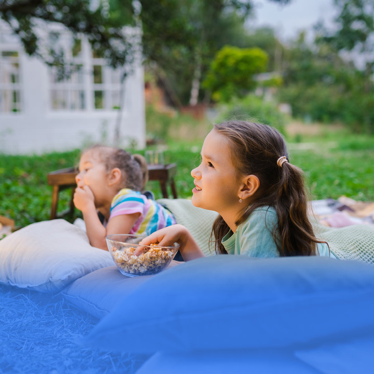 Two young girls watching an outdoor movie on cushions and eating popcorn in a garden